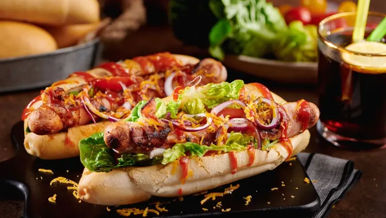 BBQ-hotdog | Venclíkovi.cz Dva grilované hotdogy v dlouhých houskách, bohatě obložené čerstvou zeleninou, červenou cibulí a přelité omáčkou, servírované na černém talíři. Vedle hotdogů je sklenice s nápojem a citronovým plátkem, a v pozadí jsou vidět další přísady a zelenina.