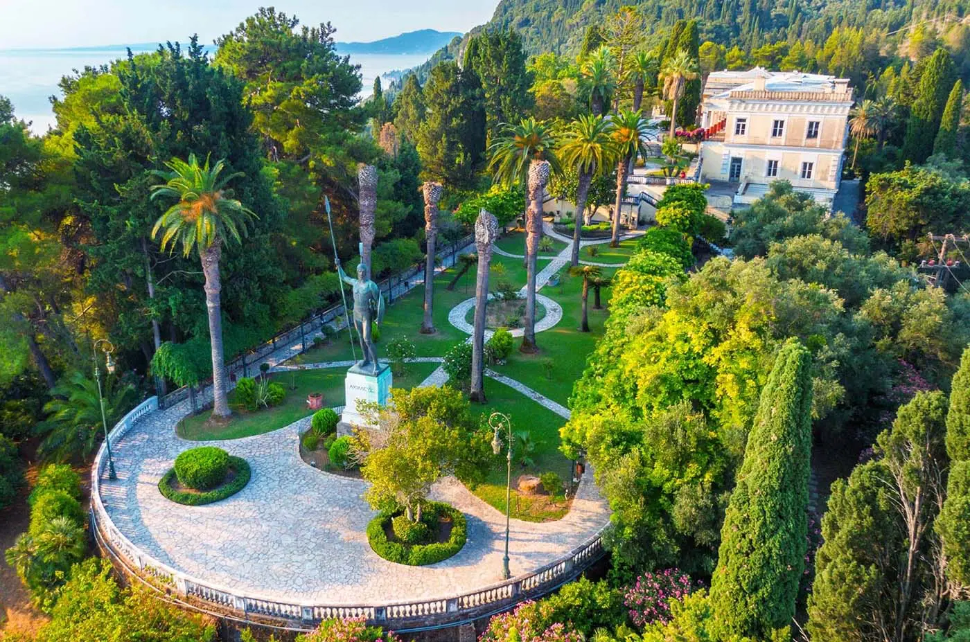 achillion korfu | Venclíkovi.cz Zahrady paláce Achillion na Korfu s sochou Achilla obklopenou zelení a palmami, v pozadí elegantní palác.