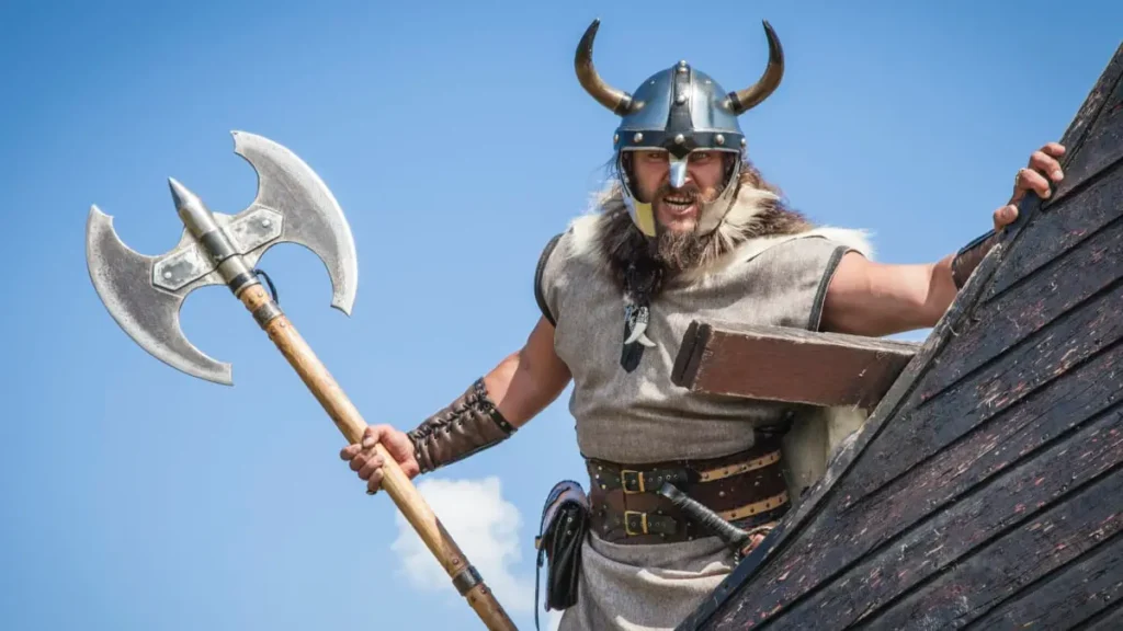 Muž v kostýmu Vikinga s rohatou helmou a sekerou pózuje na vikingské lodi – ilustrační ukázka největšího mýtu o Vikinzích, kteří ve skutečnosti helmy s rohy nikdy nenosili.