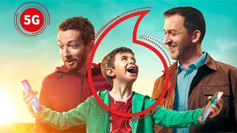 Reklamní obrázek Vodafone zobrazující tři usměvavé osoby – chlapce uprostřed a dva dospělé muže po jeho stranách – držící mobilní telefony. V levém horním rohu je logo 5G a přes střed je grafický prvek ve tvaru loga Vodafone.
