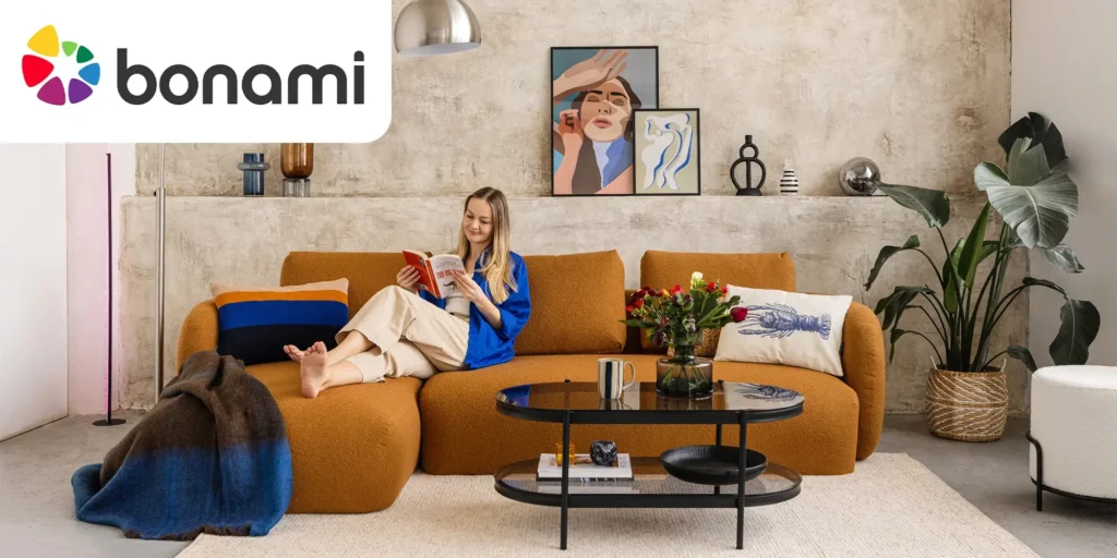 Žena odpočívá na hořčicově oranžové pohovce v moderním obývacím pokoji zařízeném ve stylu Bonami; vedle ní květiny, dekorační polštáře a minimalistický nábytek na betonové stěně s uměleckými obrazy.