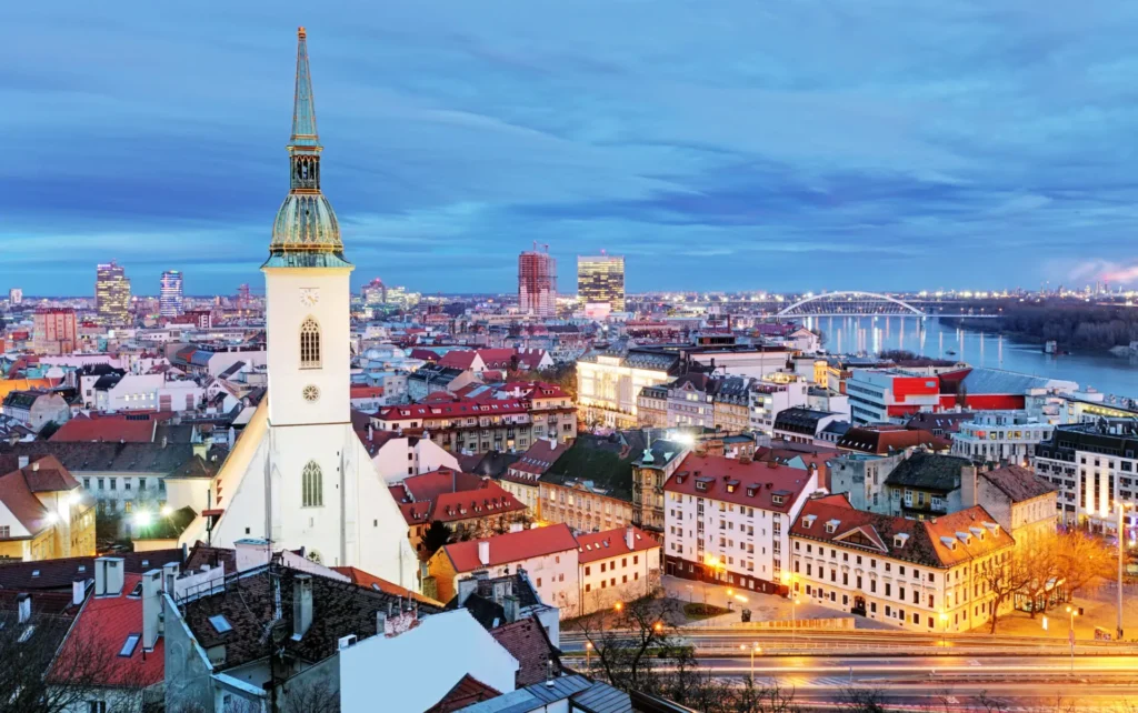 Panoramatický pohled na centrum Bratislavy s dominantou Dómu sv. Martina, Dunajem a moderními budovami v pozadí při večerním osvětlení – typická městská scenérie hlavního města Slovenska.