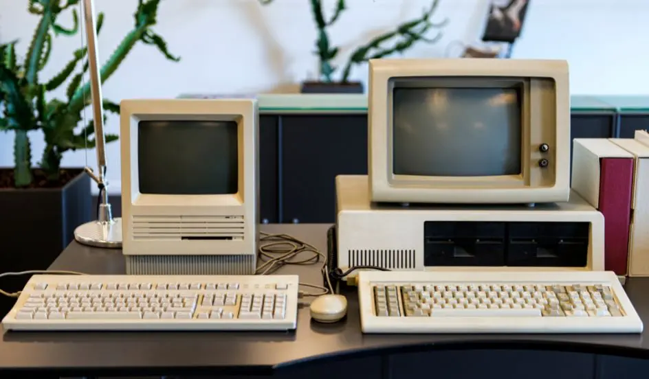 Historické stolní počítače – vlevo kompaktní Macintosh s integrovaným monitorem a disketovou mechanikou, vpravo klasický IBM PC model 5150 s dvoudisketovou jednotkou, klávesnicí a starým CRT monitorem.