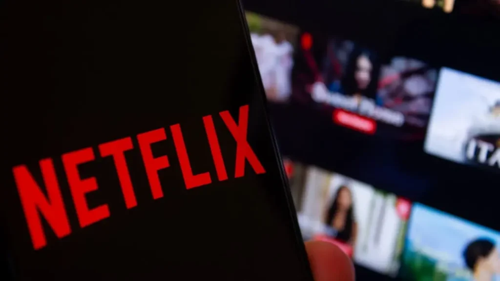 Detailní záběr loga Netflix na mobilu, v pozadí rozmazaná obrazovka s nabídkou filmů – ilustruje sledování Netflixu na cestách nebo mimo domov.