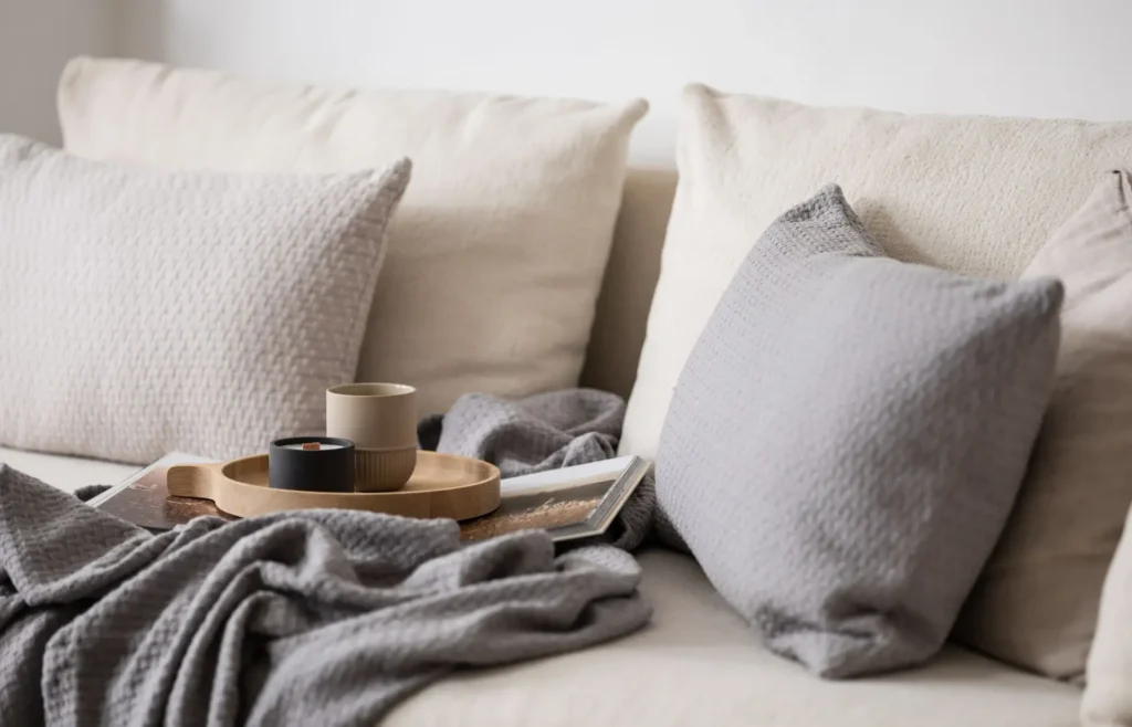 Pohodlný krémový gauč s šedými polštáři a dekou v podzimním hygge stylu, doplněný dřevěným tácem s hrnkem a svíčkou, ideální pro klidný večer doma.