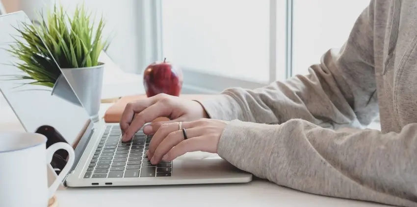 Osoba v mikině pracuje z domova na notebooku u stolu s květinou, jablkem a šálkem, typické pracovní prostředí pro homeoffice.