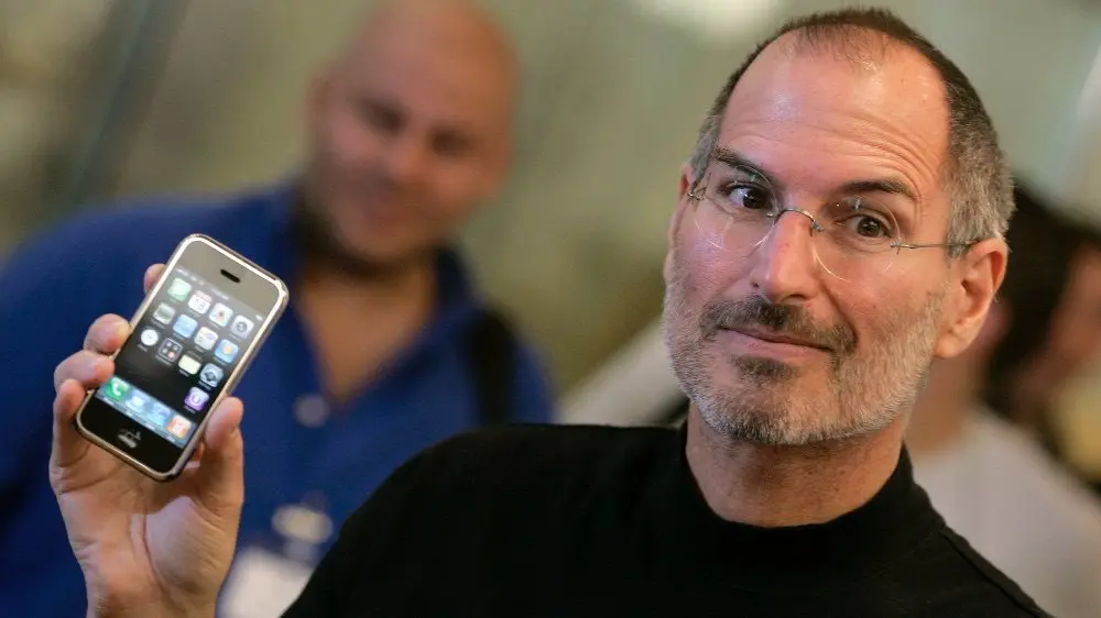 Steve Jobs drží v ruce první generaci iPhonu během jeho představení – symbol začátku éry moderních chytrých telefonů a revoluce v mobilních technologiích.