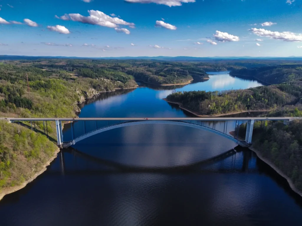 Letecký pohled na Žďákovský most, který elegantně překlenuje modrou hladinu Orlické přehrady, obklopenou lesnatou krajinou a rozlehlými kopci pod modrou oblohou s bílými mraky.