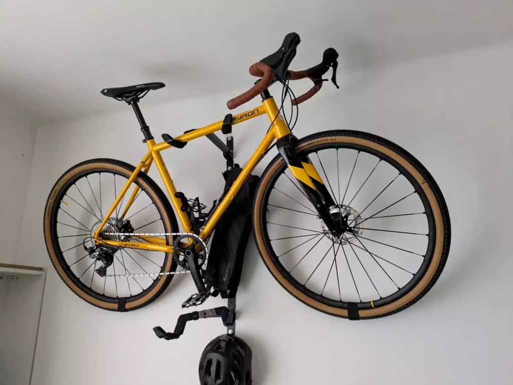 Zazimované žluté gravel kolo zavěšené na zdi v bytě, připravené na zimu a chráněné před poškozením, pod ním cyklistická helma.