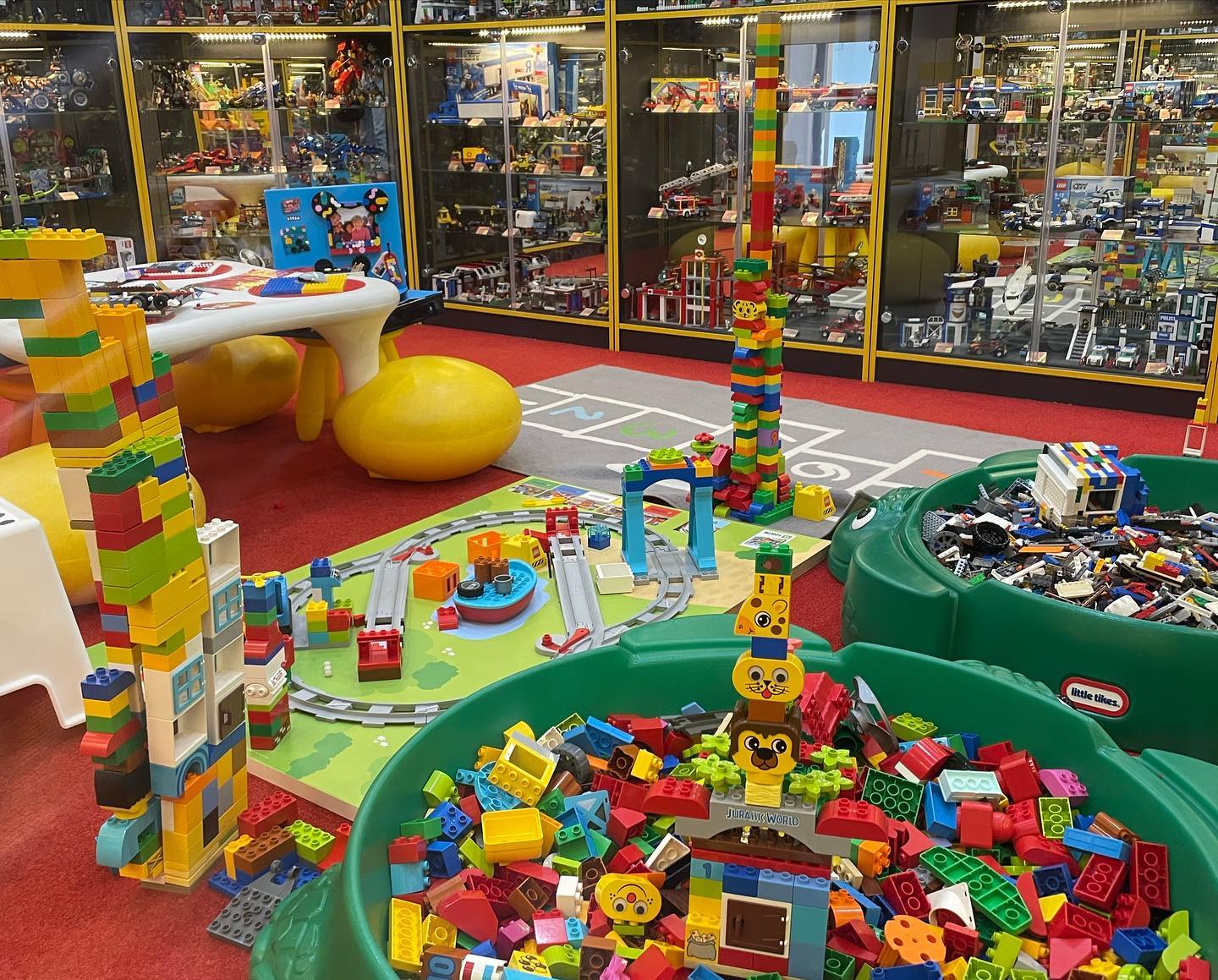 Barevný dětský koutek v Museum of Bricks s LEGO DUPLO kostkami, vysokými stavbami, vláčky a herními stoly v pozadí. 