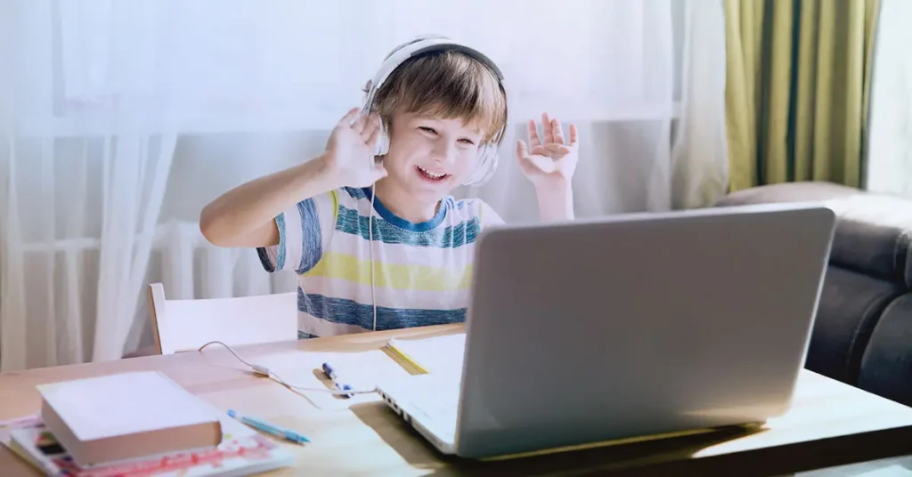 „Usměvavý kluk s velkými sluchátky sedí u notebooku, mává do videohovoru a užívá si online komunikaci z domova.