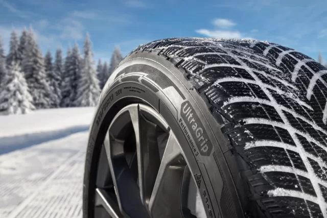 Detail zimní pneumatiky Goodyear UltraGrip Performance 3 na zasněžené silnici s lesem v pozadí.