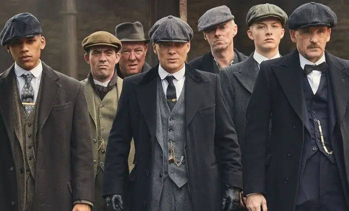 Postavy ze seriálu Peaky Blinders kráčí ulicí Birminghamu v elegantních oblecích a čepicích