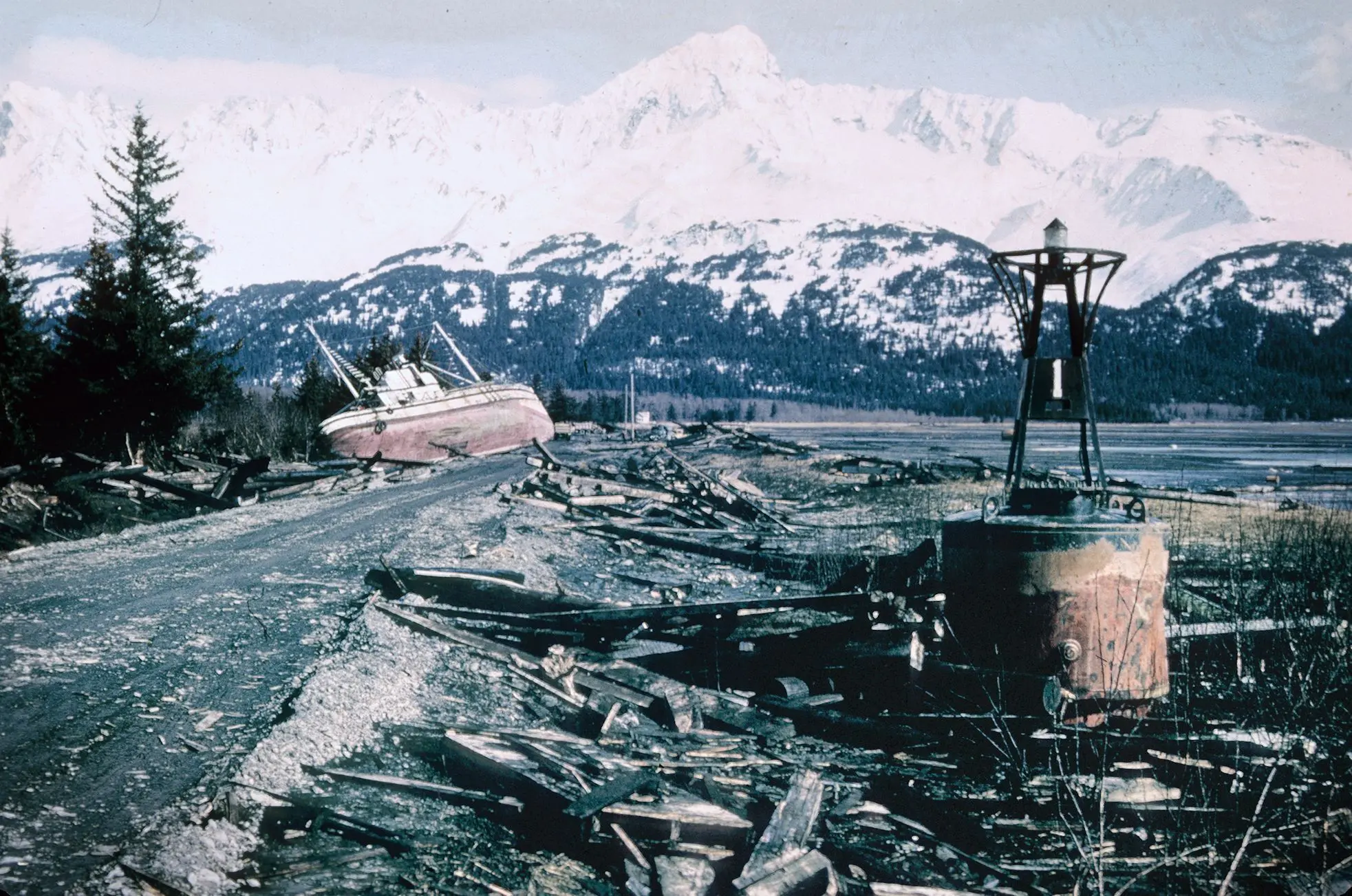 Loď vyhozená na břeh po zemětřesení u Krysích ostrovů na Aljašce. Všude trosky dřeva a v pozadí zasněžené hory, které kontrastují s tím, co zbylo po katastrofě.