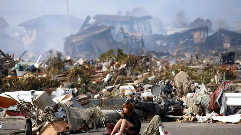 Mladá žena sedí uprostřed trosek po obřím zemětřesení v Tóhoku. Všude kolem rozbité domy, zkáza a ticho po katastrofě.