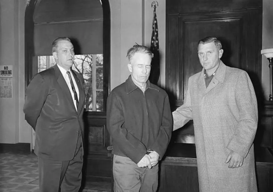 Ed Gein stojí se sklopenou hlavou u soudu, doprovázen dvěma muži z policejního doprovodu během přelíčení ve Wisconsinu.