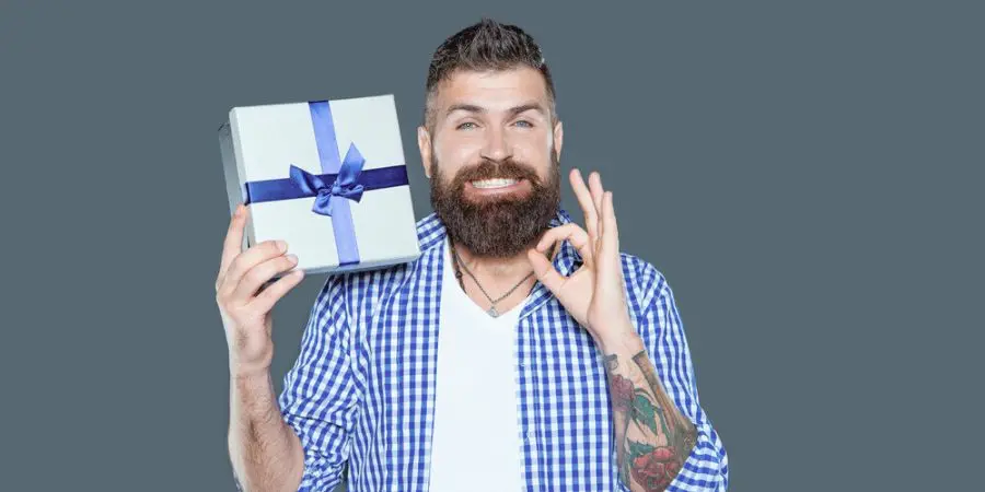 Muž s plnovousem drží dárkovou krabičku s modrou stuhou a spokojeně ukazuje gesto „OK“.