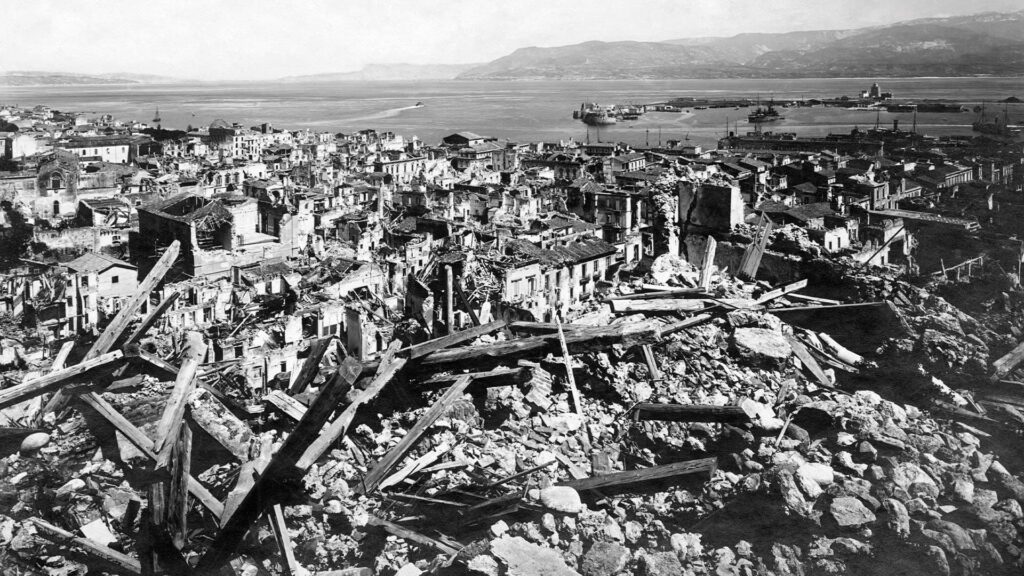 Pohled na zdevastované město Messina na Sicílii po ničivém zemětřesení a následné tsunami v roce 1908 – trosky domů, zničené ulice a panorama přístavu v pozadí.