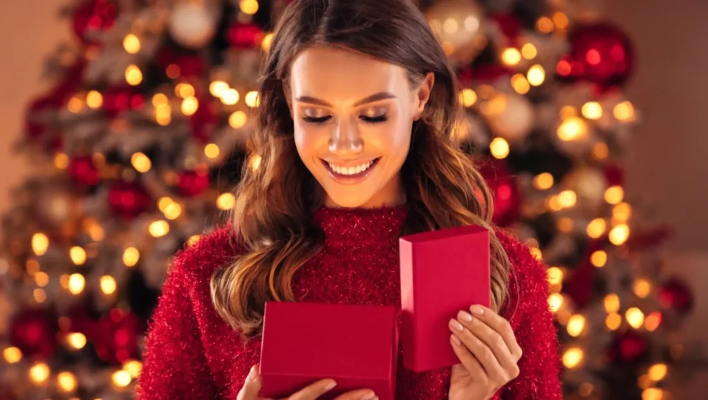 Usměvavá žena v červeném svetru otevírá dárek u vánočního stromku