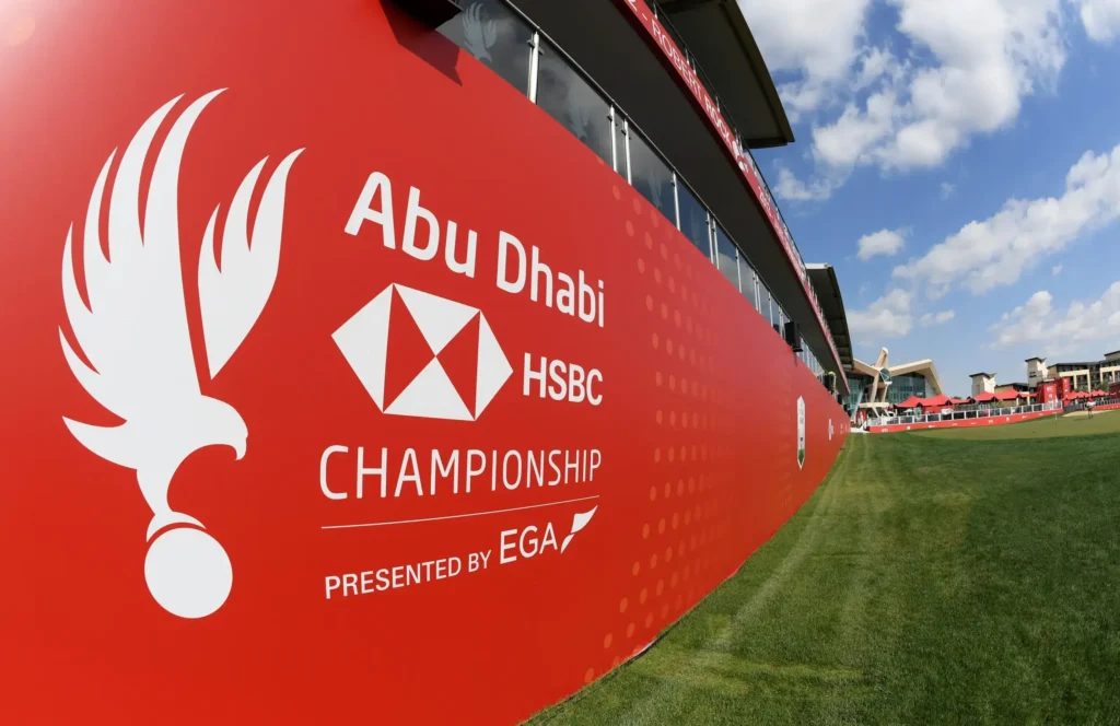 Velká červená plocha s logem Abu Dhabi HSBC Championship na golfovém hřišti a za tím jsou tribuny a krásné počasí