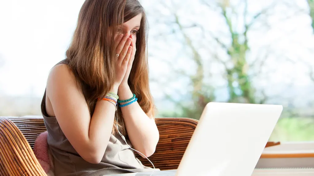 Dívka vyděšeně kouká do notebooku po nepříjemném online zážitku nebo kyberšikaně.