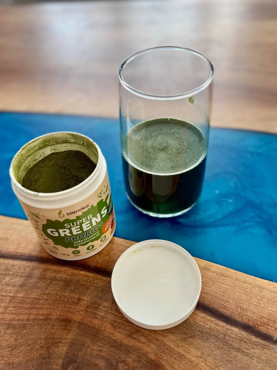 Rozmíchaný zelený nápoj Supergreens Probio Mango ve sklenici, vedle otevřená dóza s práškem