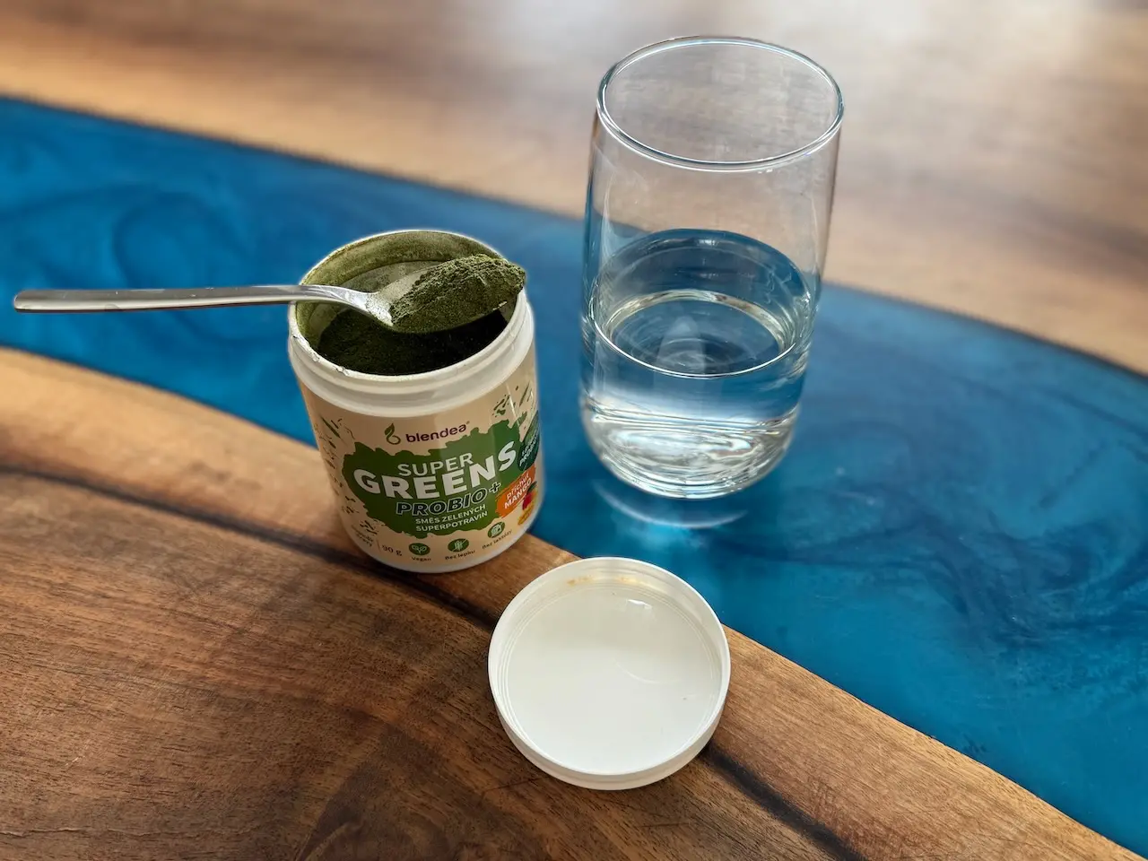 Otevřený Supergreens Probio Mango, plná lžička zeleného prášku a sklenice vody vedle