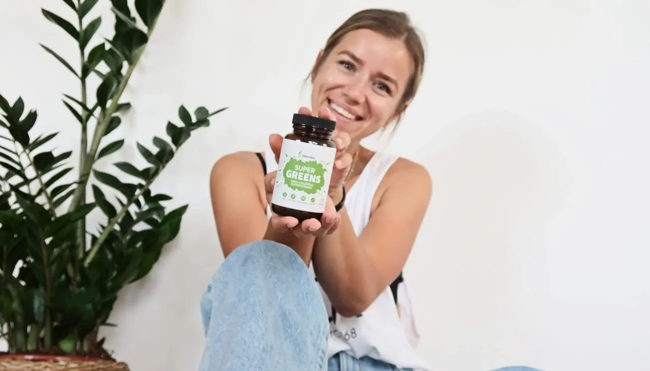 Usměvavá žena ukazuje balení Blendea Supergreens a sedí vedle zelené rostliny