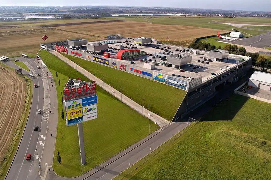 OC Šestka | Venclíkovi.cz Letecký pohled na nákupní centrum Šestka s travnatými stěnami, parkovištěm na střeše a ranvejí letiště v pozadí.