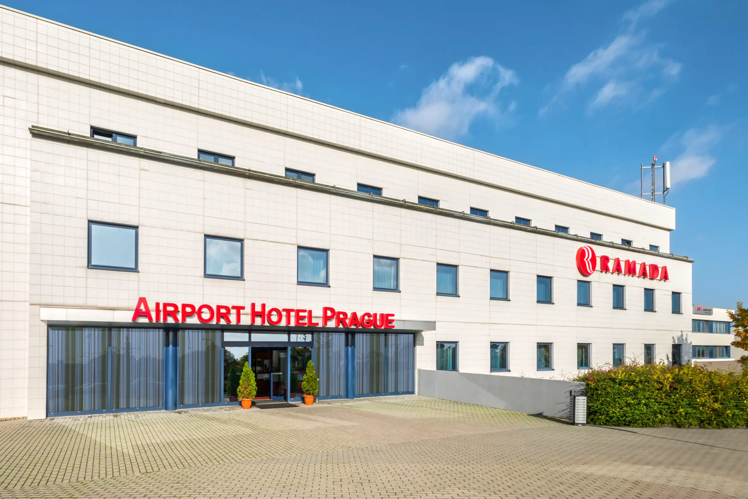 Ramada by Wyndham Airport Prague | Venclíkovi.cz Čistá bílá fasáda hotelu Ramada (Airport Hotel Prague) s červeným nápisem a dlážděným parkovištěm před vchodem.