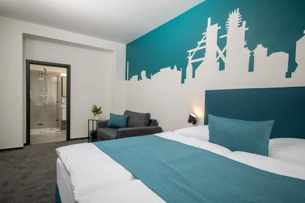 VP1 Ostrava 5 | Venclíkovi.cz Moderní světlý pokoj v Hotelu VP1 s výraznou tyrkysovou stěnou a bílou siluetou industriálních věží.