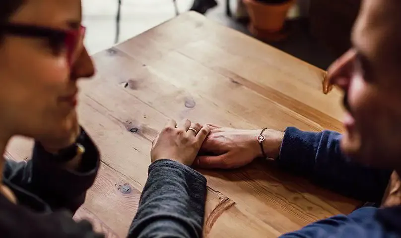 komunikace mimo loznici | Venclíkovi.cz Pár sedí u stolu, drží se za ruce a vede vážný, ale klidný rozhovor.