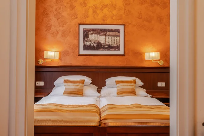 Dvoulůžkový pokoj hotelu Radium Palace se zlatavými textiliemi a oranžovou stěnou