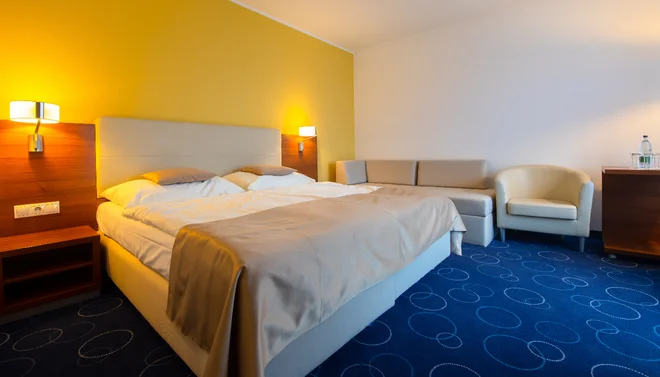 Dvoulůžkový pokoj hotelu Panorama se žlutou stěnou, modrým kobercem a posezením