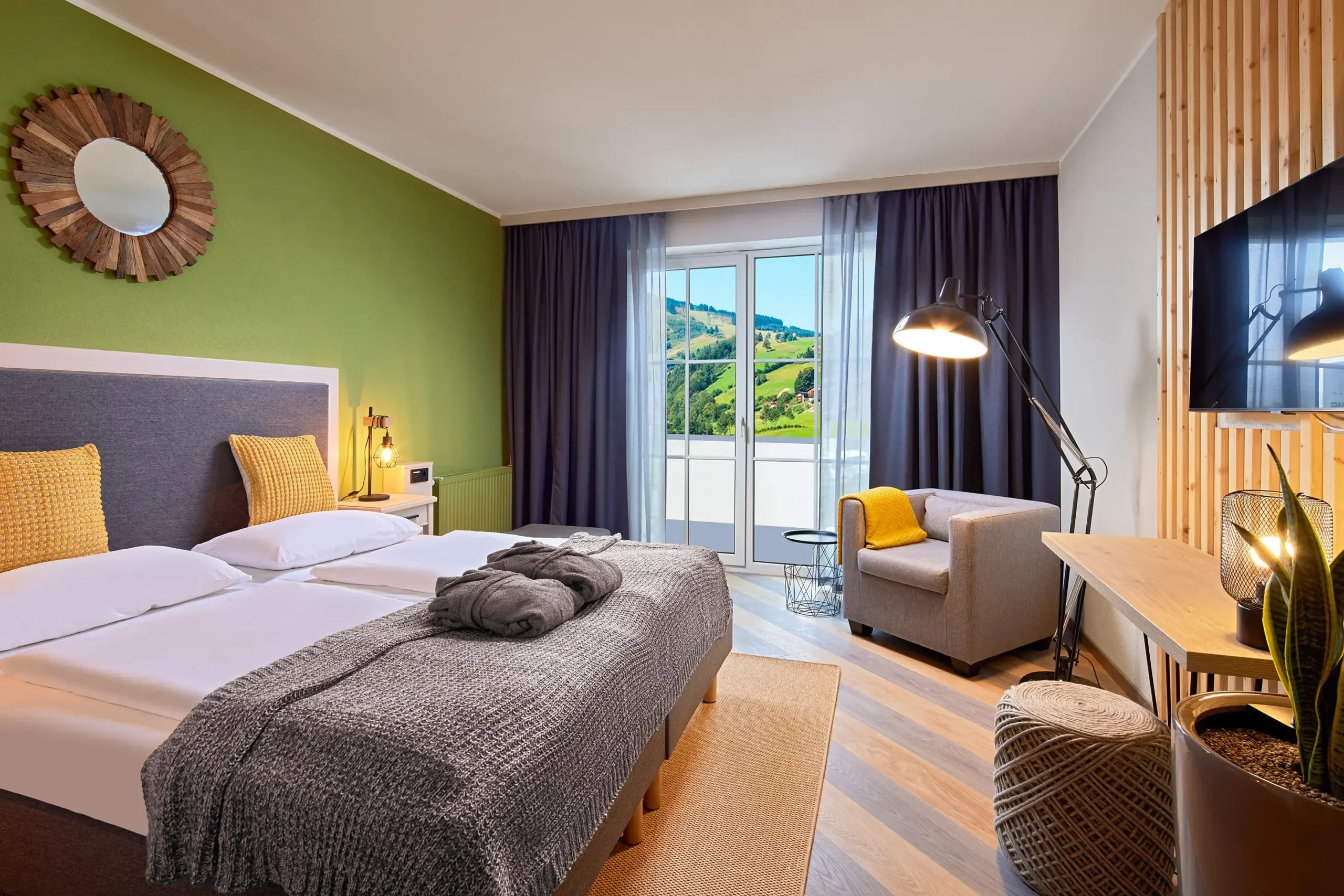 Světle laděný pokoj hotelu ever.grün Kaprun s manželskou postelí, posezením a balkonem s výhledem na zelené kopce Kaprunu.